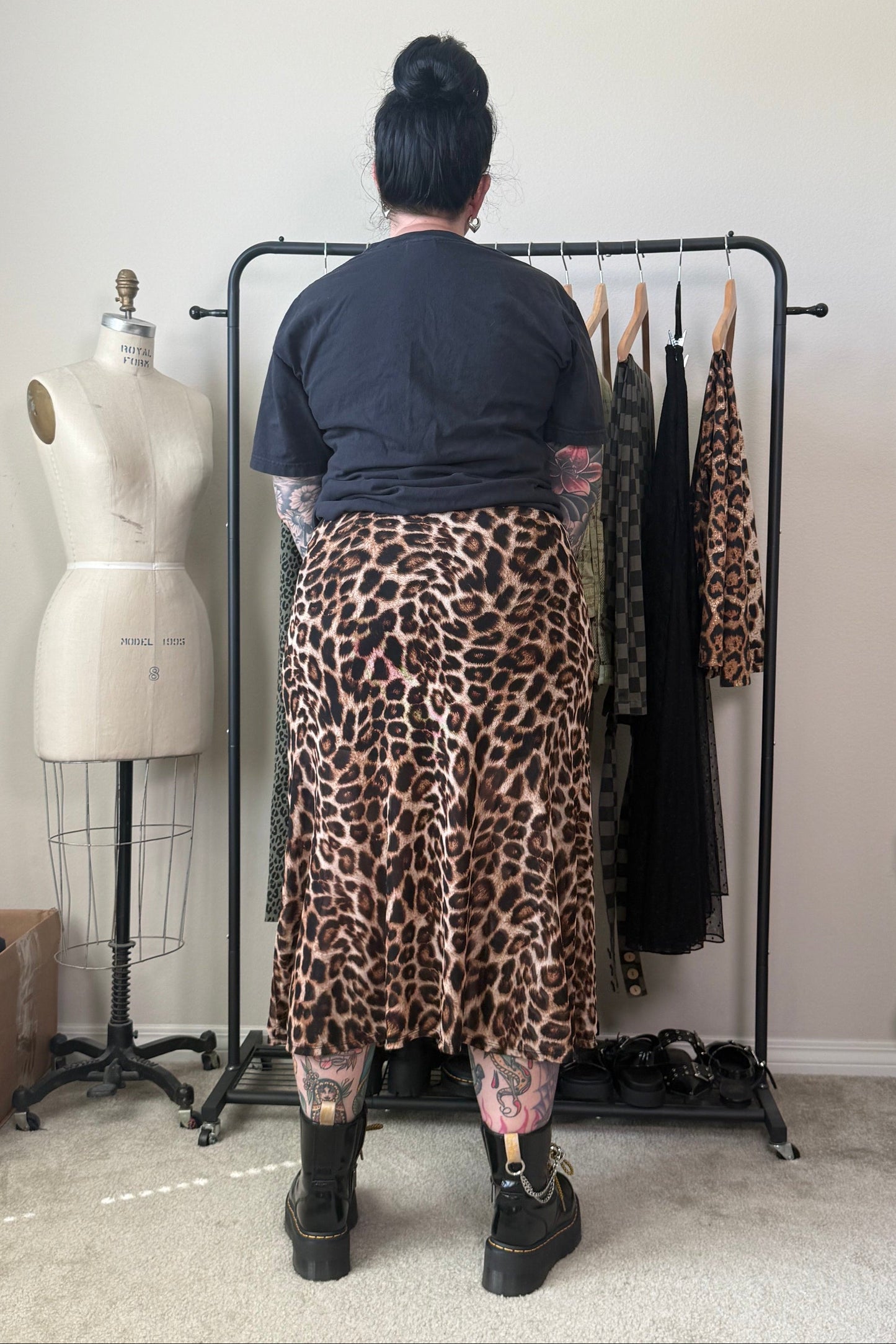 A person stands with their back to the camera wearing a leopard-print midi skirt and a dark t-shirt, standing in front of a clothing rack in a room with a dress form and shelves.