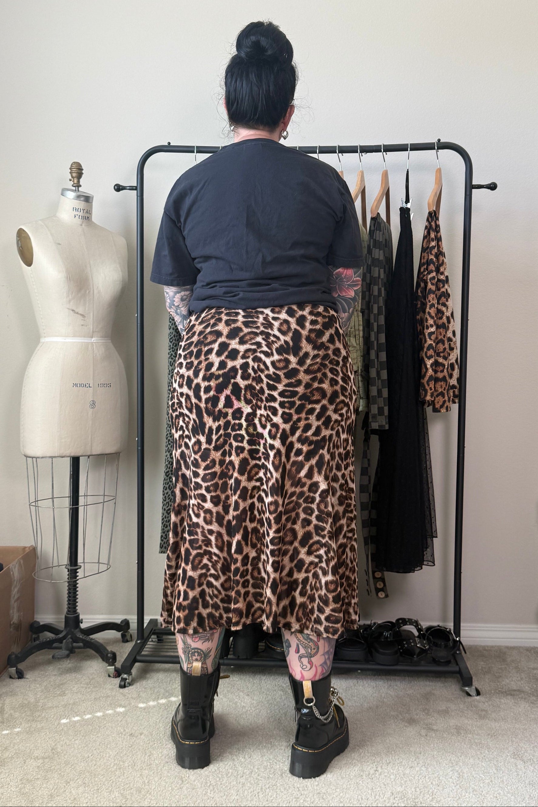 A person stands with their back to the camera wearing a leopard-print midi skirt and a dark t-shirt, standing in front of a clothing rack in a room with a dress form and shelves.