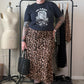 A person stands in a room wearing a black graphic t-shirt, a leopard print midi skirt, black combat boots, and holding a black woven handbag. A clothing rack with other garments, a dress form, and a beige wall are visible in the background.