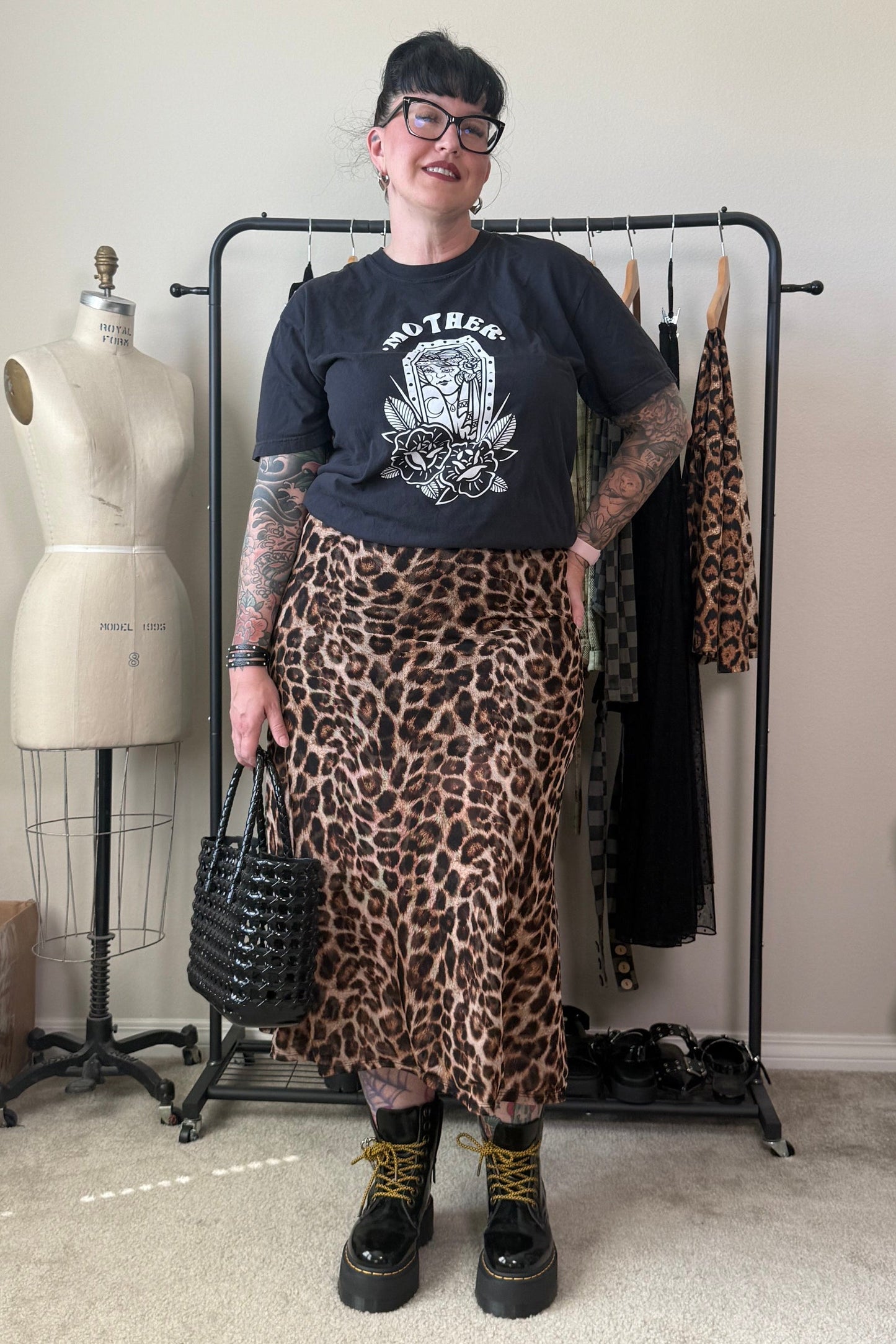 A person stands in a room wearing a black graphic t-shirt, a leopard print midi skirt, black combat boots, and holding a black woven handbag. A clothing rack with other garments, a dress form, and a beige wall are visible in the background.