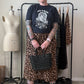 A person stands in a room wearing a black graphic t-shirt, a leopard print midi skirt, black combat boots with yellow laces, and holding a black studded handbag. A dress form and clothing rack are visible in the background.
