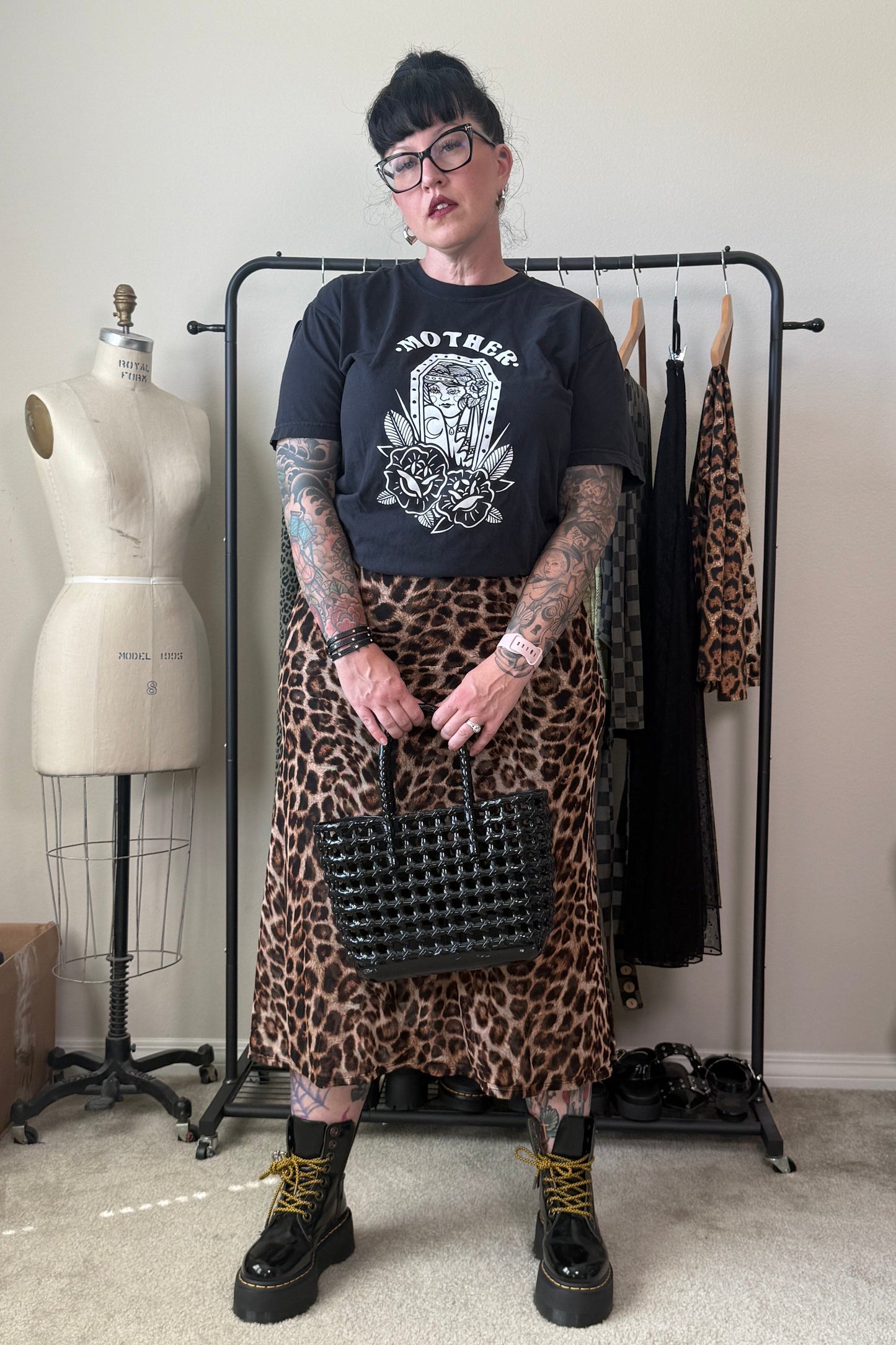 A person stands in a room wearing a black graphic t-shirt, a leopard print midi skirt, black combat boots with yellow laces, and holding a black studded handbag. A dress form and clothing rack are visible in the background.