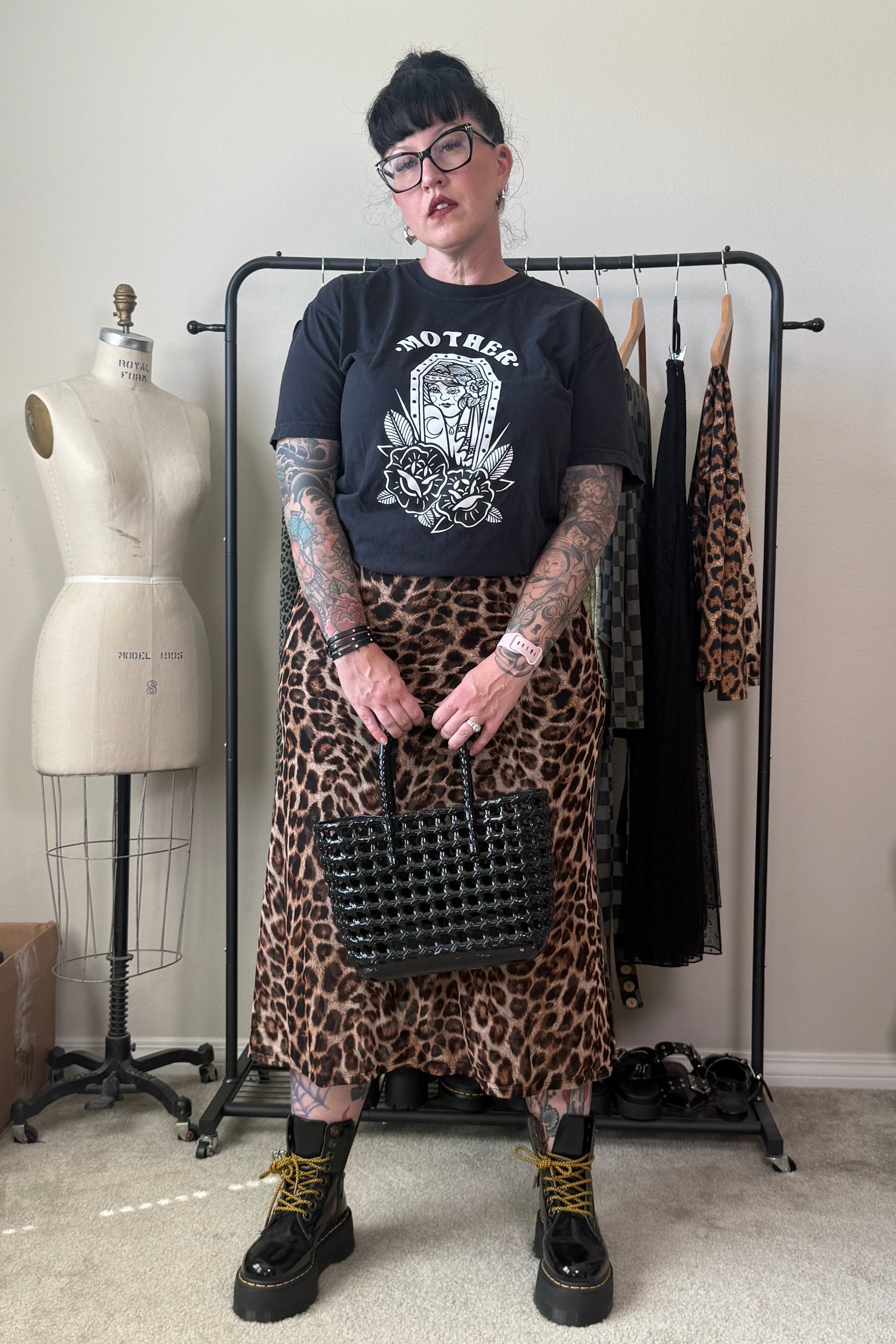 A person stands in a room wearing a black graphic t-shirt, a leopard print midi skirt, black combat boots with yellow laces, and holding a black studded handbag. A dress form and clothing rack are visible in the background.