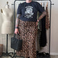 A person stands indoors wearing a black graphic T-shirt, a leopard-print midi skirt, black boots, and holding a black woven handbag. A clothing rack with garments, a dress form, and room elements are visible in the background.