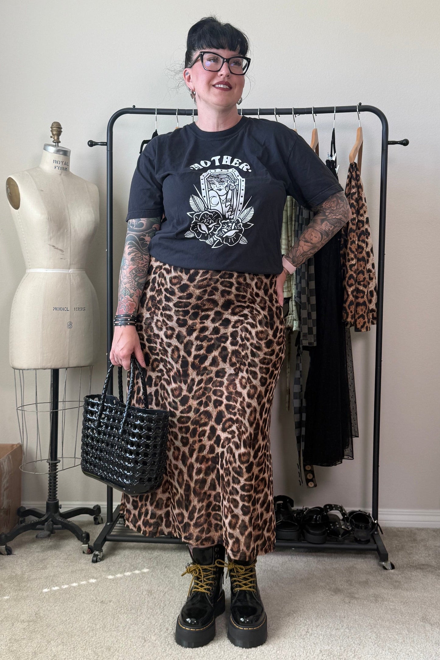 A person stands indoors wearing a black graphic T-shirt, a leopard-print midi skirt, black boots, and holding a black woven handbag. A clothing rack with garments, a dress form, and room elements are visible in the background.