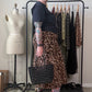 A woman with glasses and tattoos stands in a room wearing a leopard-print skirt, a black t-shirt, and black boots, holding a black woven handbag. A clothing rack with other garments, a dress form, and storage items are visible in the background.