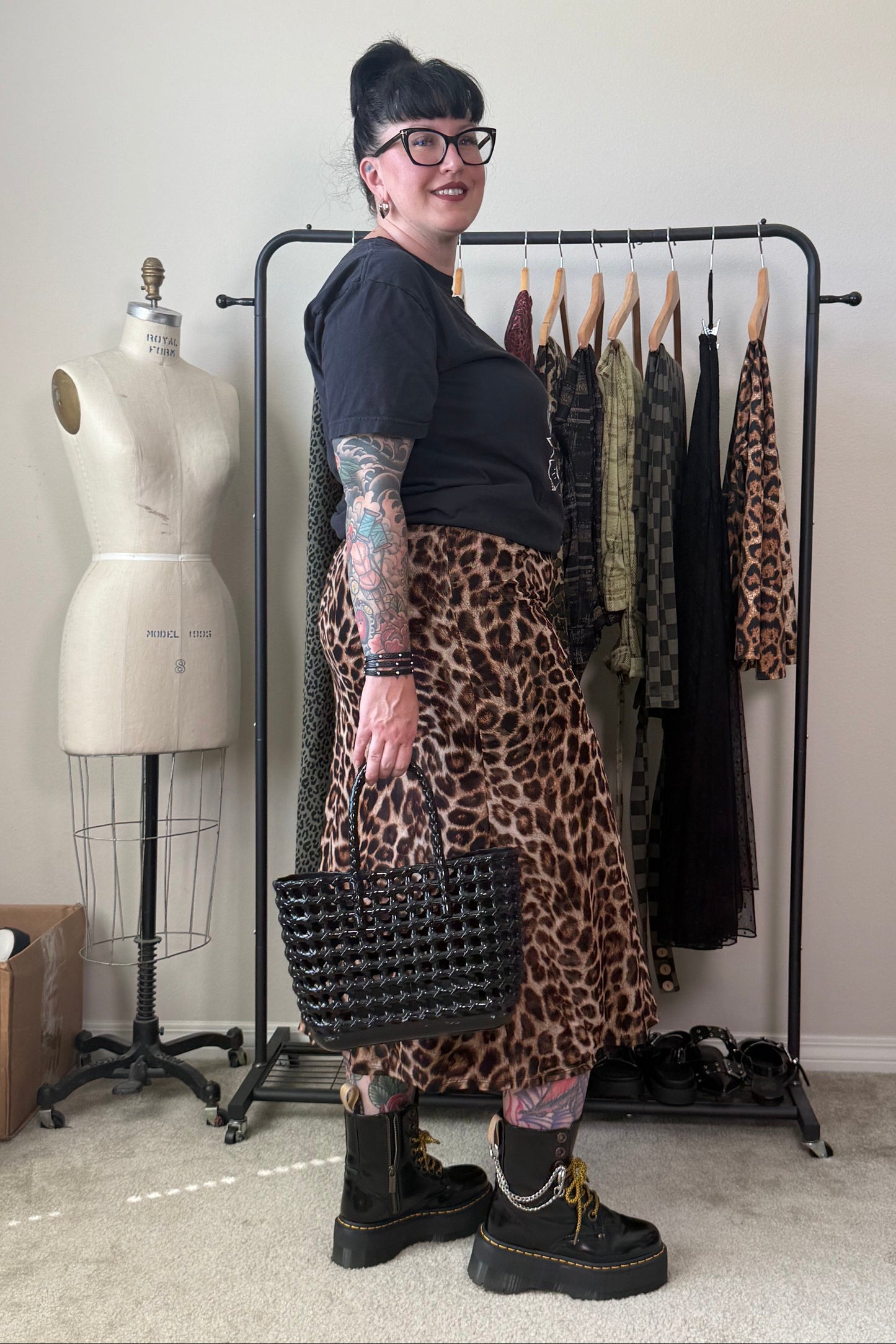 A woman with glasses and tattoos stands in a room wearing a leopard-print skirt, a black t-shirt, and black boots, holding a black woven handbag. A clothing rack with other garments, a dress form, and storage items are visible in the background.