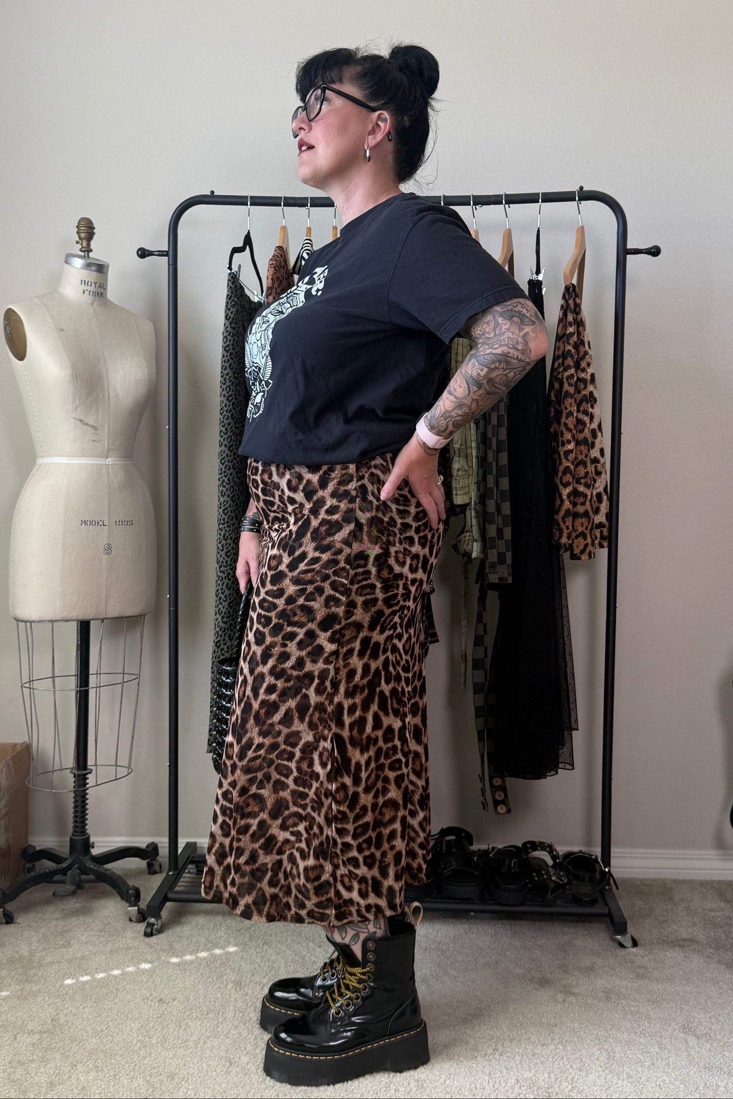 A person modeling a leopard print midi skirt with a black graphic t-shirt and black combat boots, standing in a room with a clothing rack and a dress form.