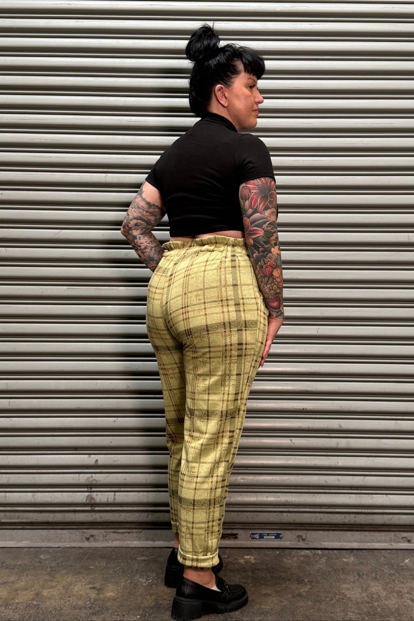 A person lying on their side in front of a gray metal roll-up door, wearing a black short-sleeve top and beige plaid pants with black shoes.