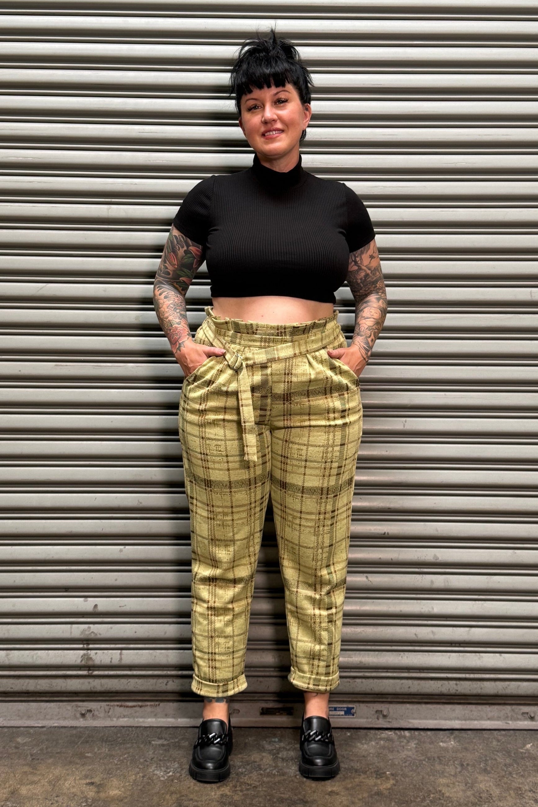 A person wearing a black cropped short-sleeve top and olive green plaid trousers, standing against a gray metal shutter door.