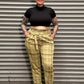 A person wearing a black cropped t-shirt and yellow plaid cargo pants stands against a gray corrugated metal shutter.