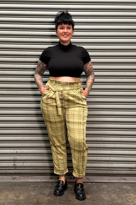A person wearing a black cropped t-shirt and yellow plaid cargo pants stands against a gray corrugated metal shutter.