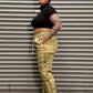 A person lying horizontally against a corrugated metal wall, wearing a black cropped turtleneck, olive green plaid trousers, and black shoes.