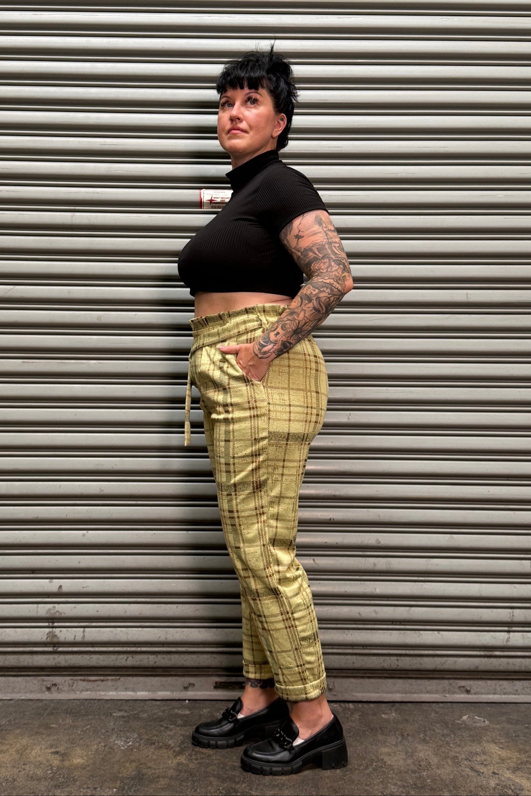 A person lying horizontally against a corrugated metal wall, wearing a black cropped turtleneck, olive green plaid trousers, and black shoes.