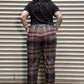 A person viewed from above wearing a black short-sleeve top and dark plaid pants with brown and purple lines, standing against a corrugated metal wall.