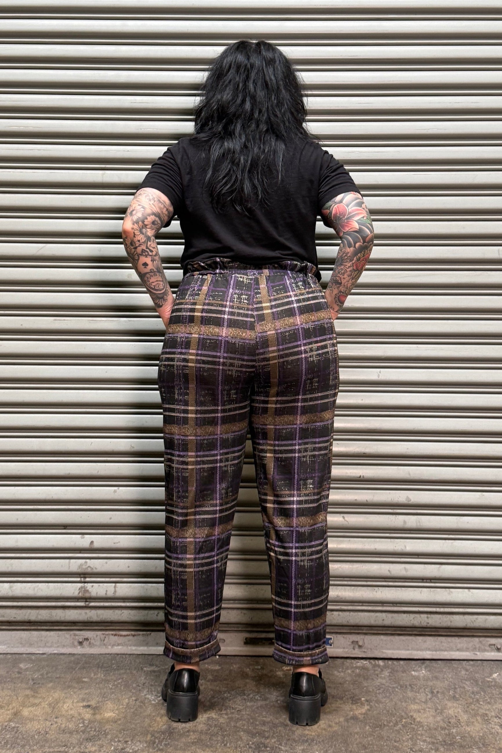 A person viewed from above wearing a black short-sleeve top and dark plaid pants with brown and purple lines, standing against a corrugated metal wall.