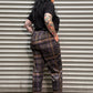 A person lying flat on their back against a gray corrugated metal shutter, wearing a black t-shirt, dark brown and black plaid pants, and black shoes.