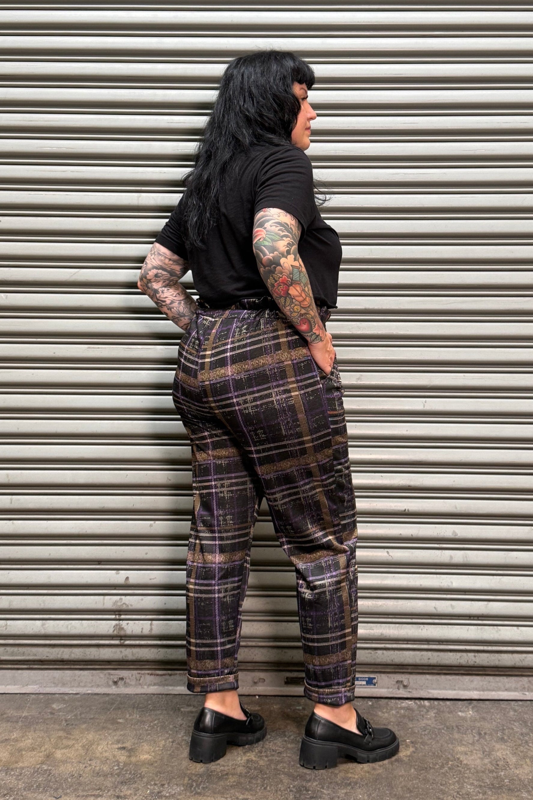 A person lying flat on their back against a gray corrugated metal shutter, wearing a black t-shirt, dark brown and black plaid pants, and black shoes.