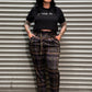 A person wearing a black graphic t-shirt and dark brown plaid pants with suspenders, standing against a gray metal roll-up door.