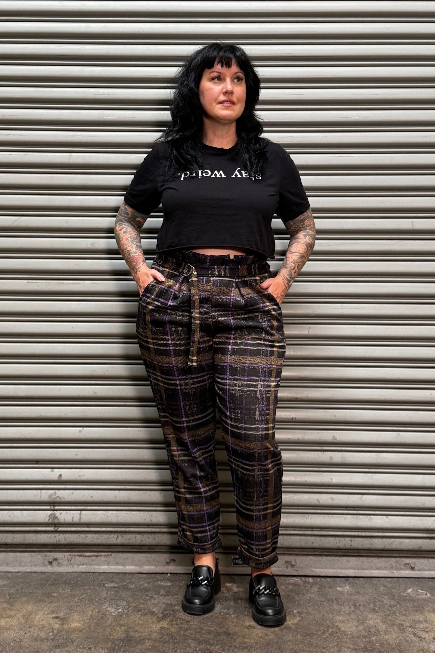 A person wearing a black graphic t-shirt and dark brown plaid pants with suspenders, standing against a gray metal roll-up door.