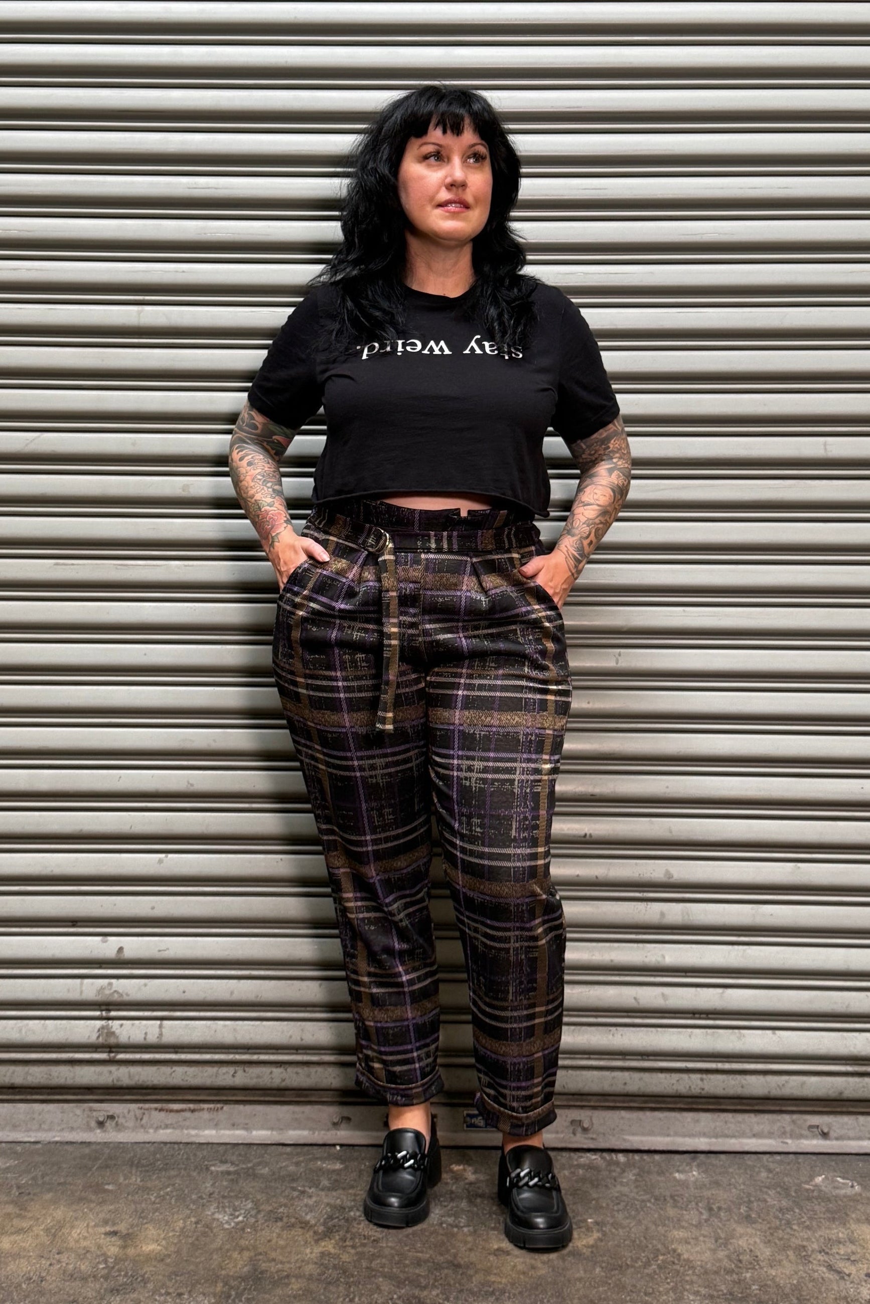 A person wearing a black graphic t-shirt and dark brown plaid pants with suspenders, standing against a gray metal roll-up door.