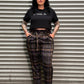 A person wearing a black cropped T-shirt with white text and dark plaid pants, standing against a corrugated metal shutter.