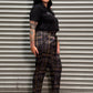 A person wearing a black short-sleeve top and dark brown plaid pants with black shoes, lying on a corrugated metal shutter against a concrete floor.