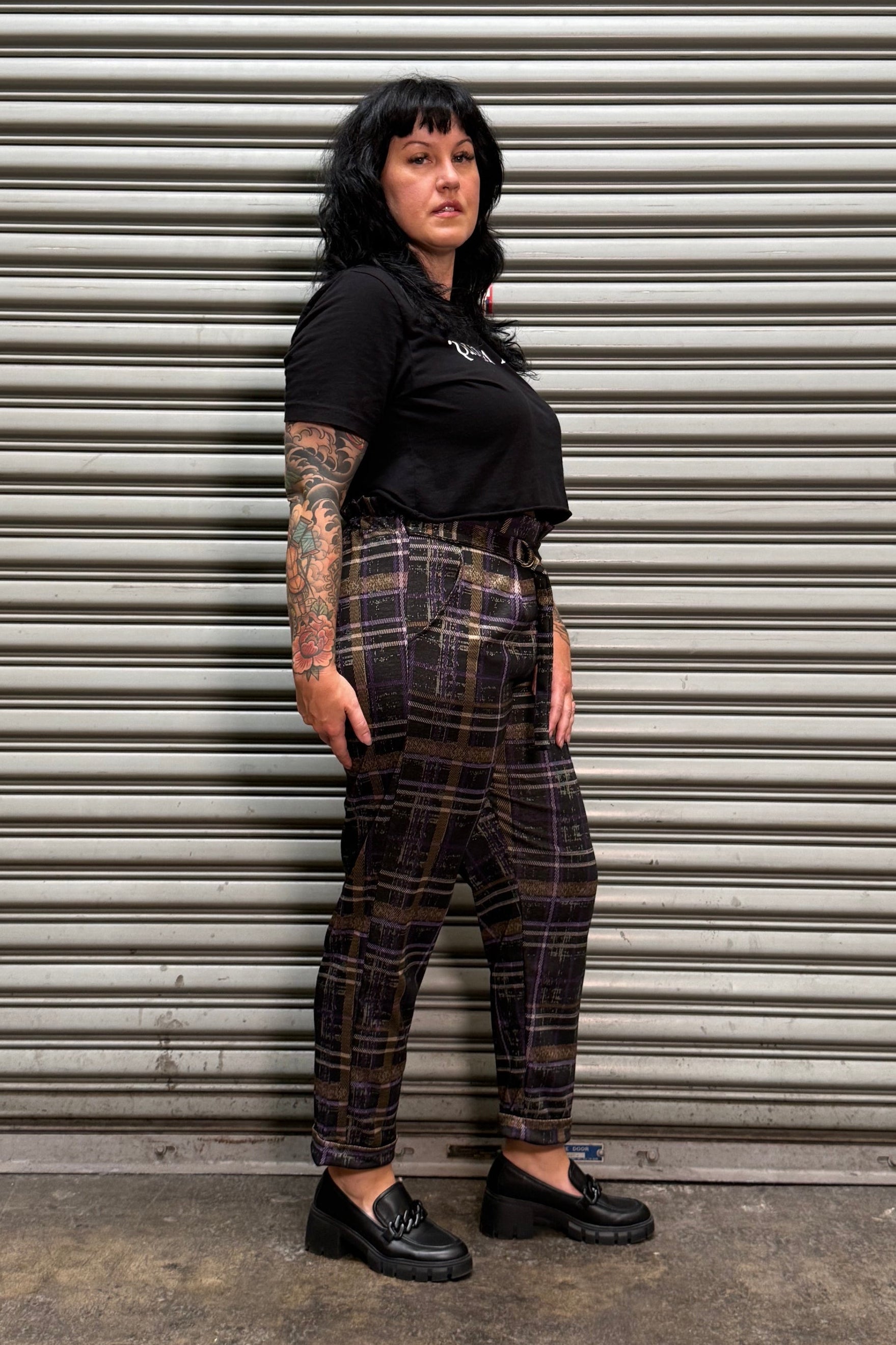 A person wearing a black short-sleeve top and dark brown plaid pants with black shoes, lying on a corrugated metal shutter against a concrete floor.