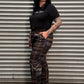 A person lying on their back against a corrugated metal garage door, wearing a black t-shirt, a brown and black plaid pair of pants, and black shoes.
