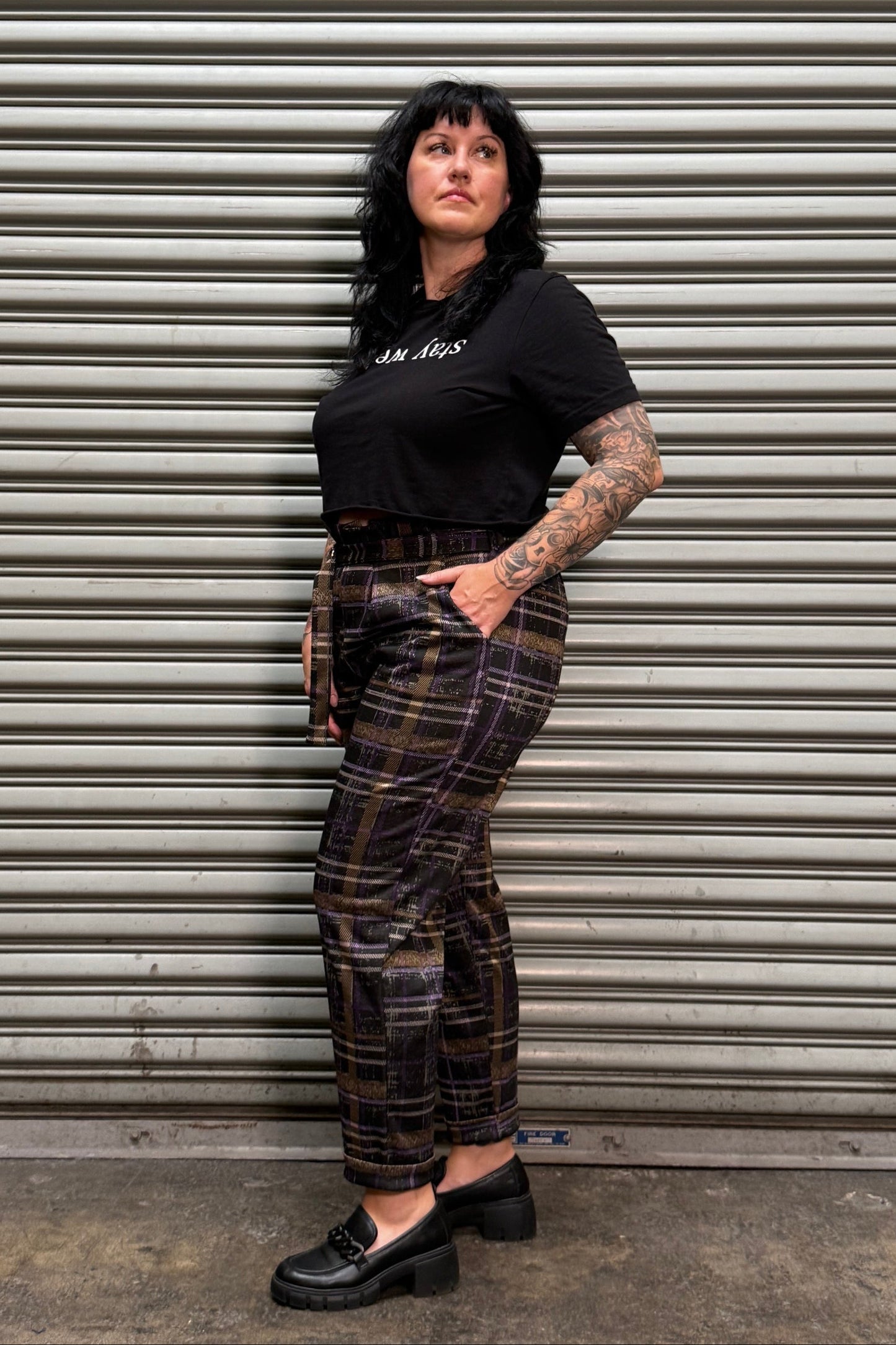 A person lying on their back against a corrugated metal garage door, wearing a black t-shirt, a brown and black plaid pair of pants, and black shoes.