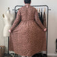 A person stands with their back to the camera wearing a brown and black leopard print long-sleeve midi dress, holding out the sides of the skirt to show the full shape. They are standing in a room with a clothing rack, a dress form, and a beige wall.