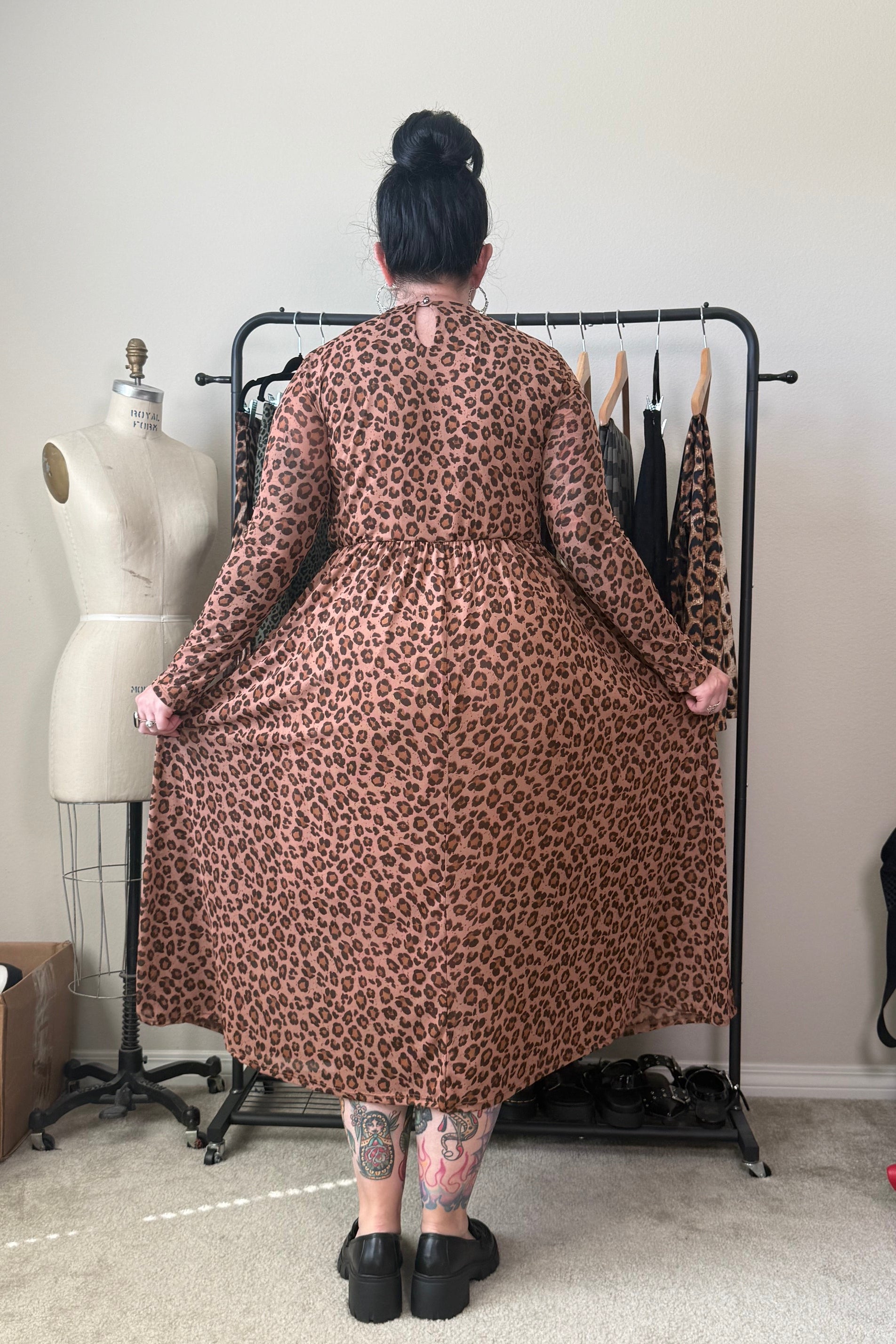A person stands with their back to the camera wearing a brown and black leopard print long-sleeve midi dress, holding out the sides of the skirt to show the full shape. They are standing in a room with a clothing rack, a dress form, and a beige wall.