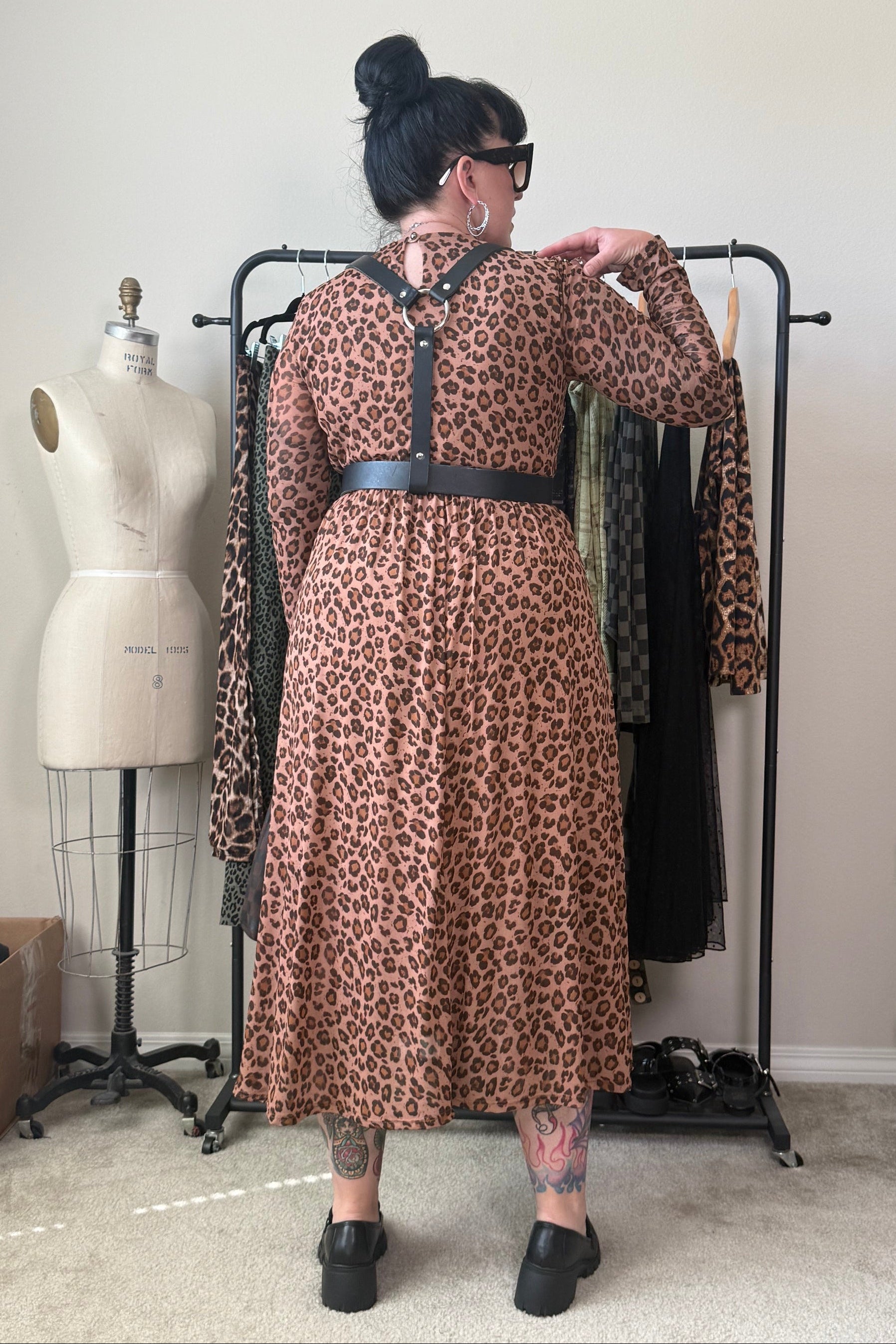 A person stands with their back to the camera wearing a leopard print midi dress with a black belt, standing in front of a clothing rack in a room with a mannequin and other clothes.