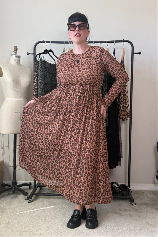 A woman wearing a brown and black leopard-print long-sleeve maxi dress stands in front of a clothing rack in a room with a mannequin and shelves.