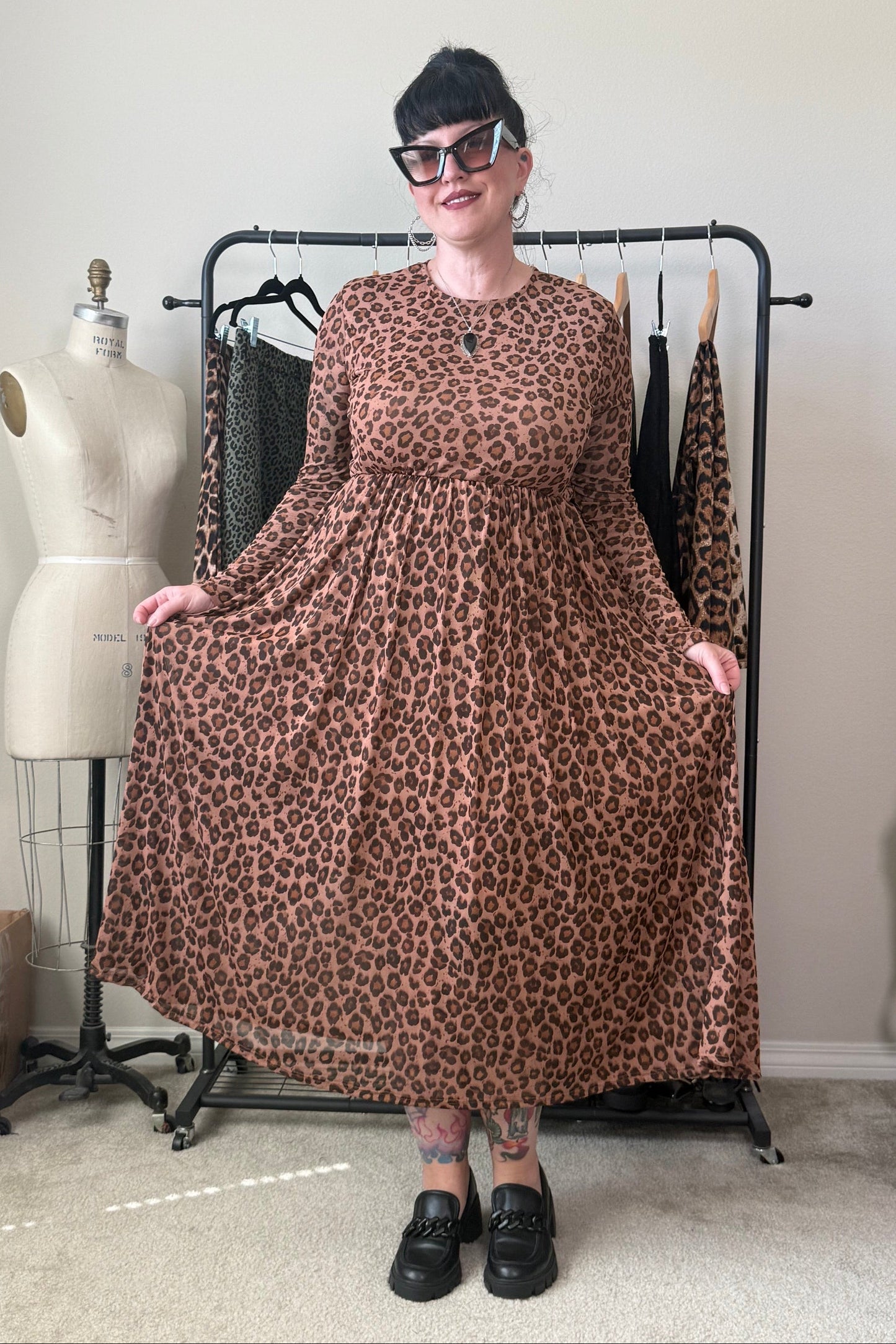 A person wearing a brown leopard-print long-sleeve maxi dress stands in front of a clothing rack in a room with a beige wall and carpet. A dress form is visible to the left.