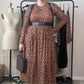 A woman stands in a room wearing a brown and black leopard-print midi dress with a wide black belt, black loafers, and holding a brown patterned handbag. A clothing rack with other garments, a dress form, and a beige wall are visible in the background.