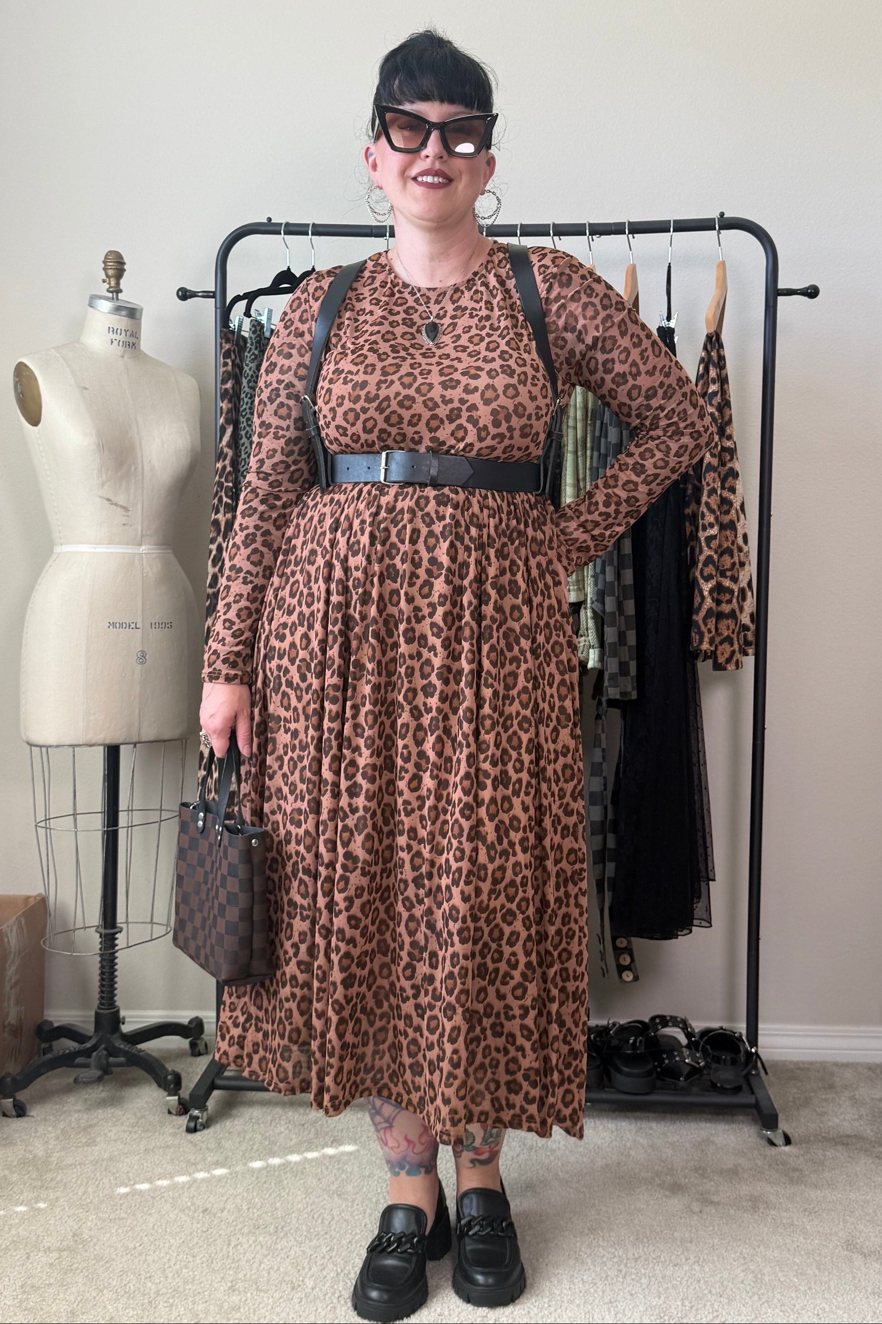 A woman stands in a room wearing a brown and black leopard-print midi dress with a wide black belt, black loafers, and holding a brown patterned handbag. A clothing rack with other garments, a dress form, and a beige wall are visible in the background.