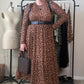 A person stands indoors wearing a brown leopard-print long-sleeve midi dress with a black belt, black shoes, and holding a patterned handbag. A clothing rack with other garments, a dress form, and room furniture are visible in the background.