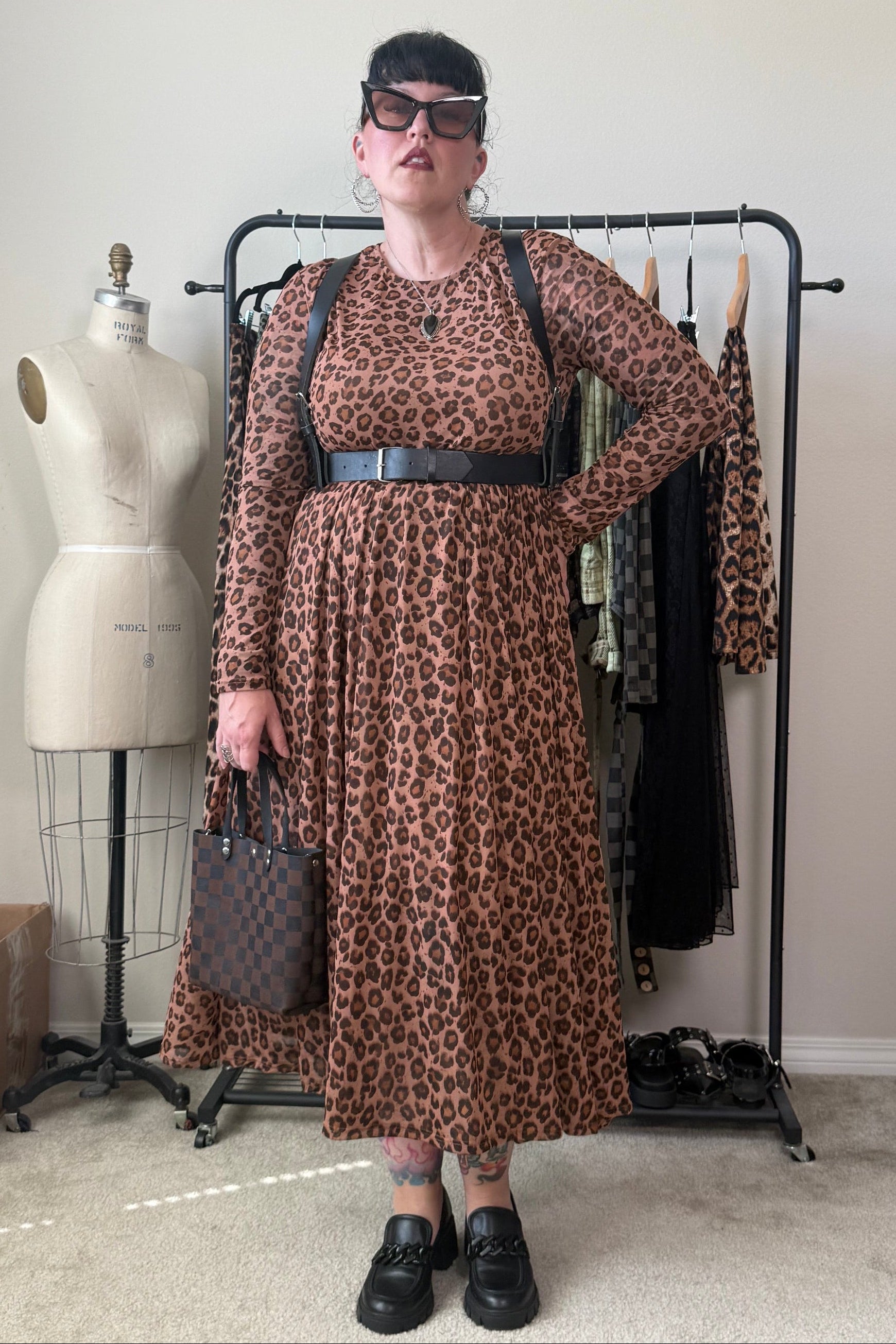 A person stands indoors wearing a brown leopard-print long-sleeve midi dress with a black belt, black shoes, and holding a patterned handbag. A clothing rack with other garments, a dress form, and room furniture are visible in the background.