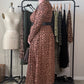 A woman wearing a brown and black leopard-print midi dress with a matching belt stands in front of a clothing rack in a room with a mannequin and other garments.