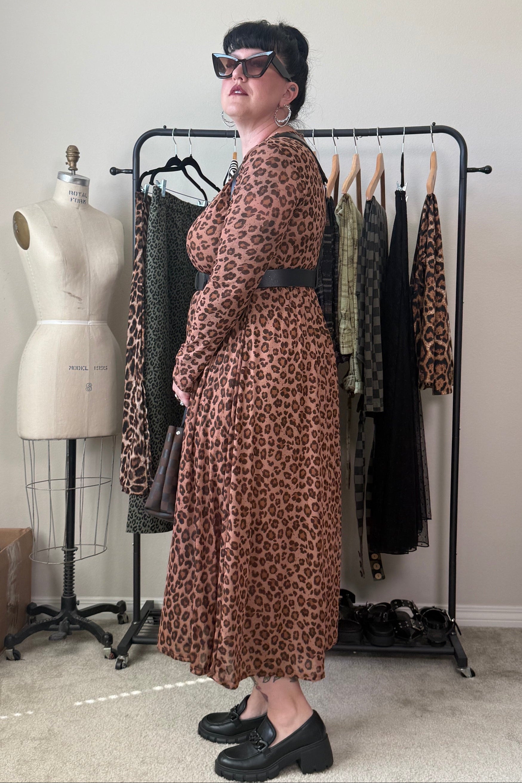 A woman wearing a brown and black leopard-print midi dress with a matching belt stands in front of a clothing rack in a room with a mannequin and other garments.
