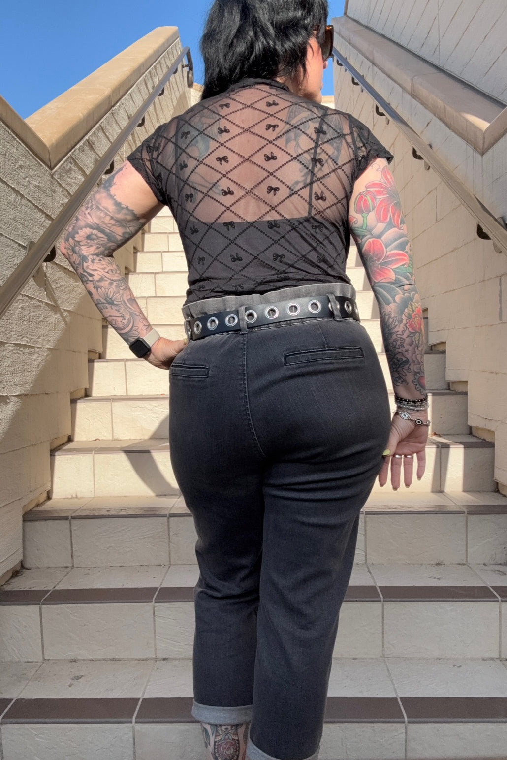Person with tattoos wearing a sheer top and dark jeans on a staircase.