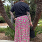Person wearing a black top and pink skirt with tattoos, standing in front of palm trees.