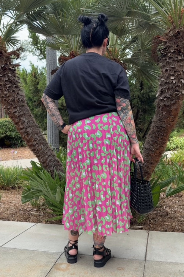 Person wearing a black top and pink skirt with tattoos, standing in front of palm trees.