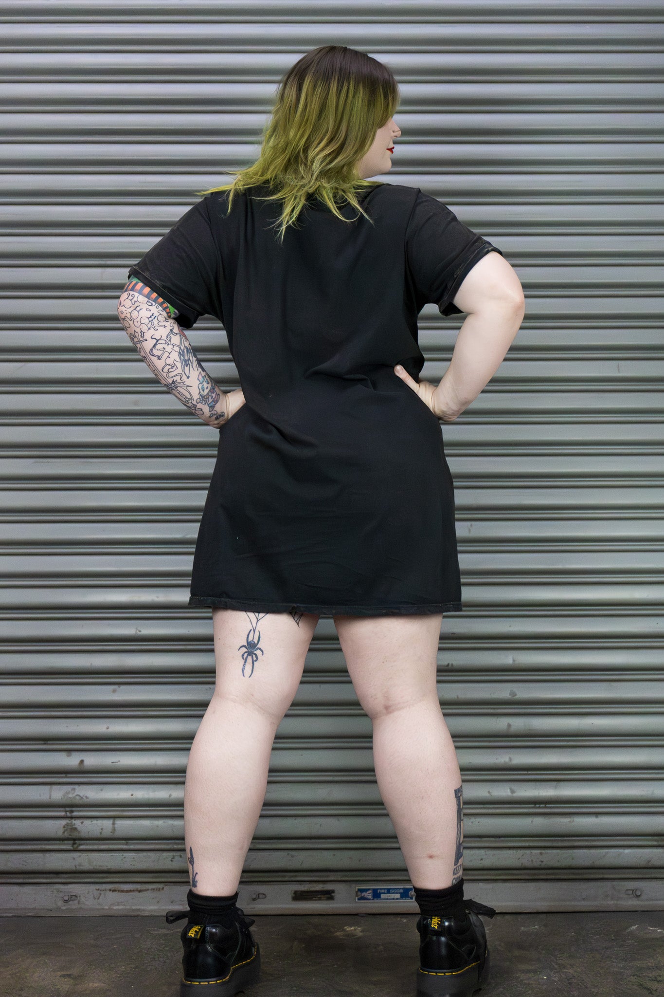 Person wearing a black t-shirt with tattoos visible on arms and legs, standing in front of a metallic shutter.