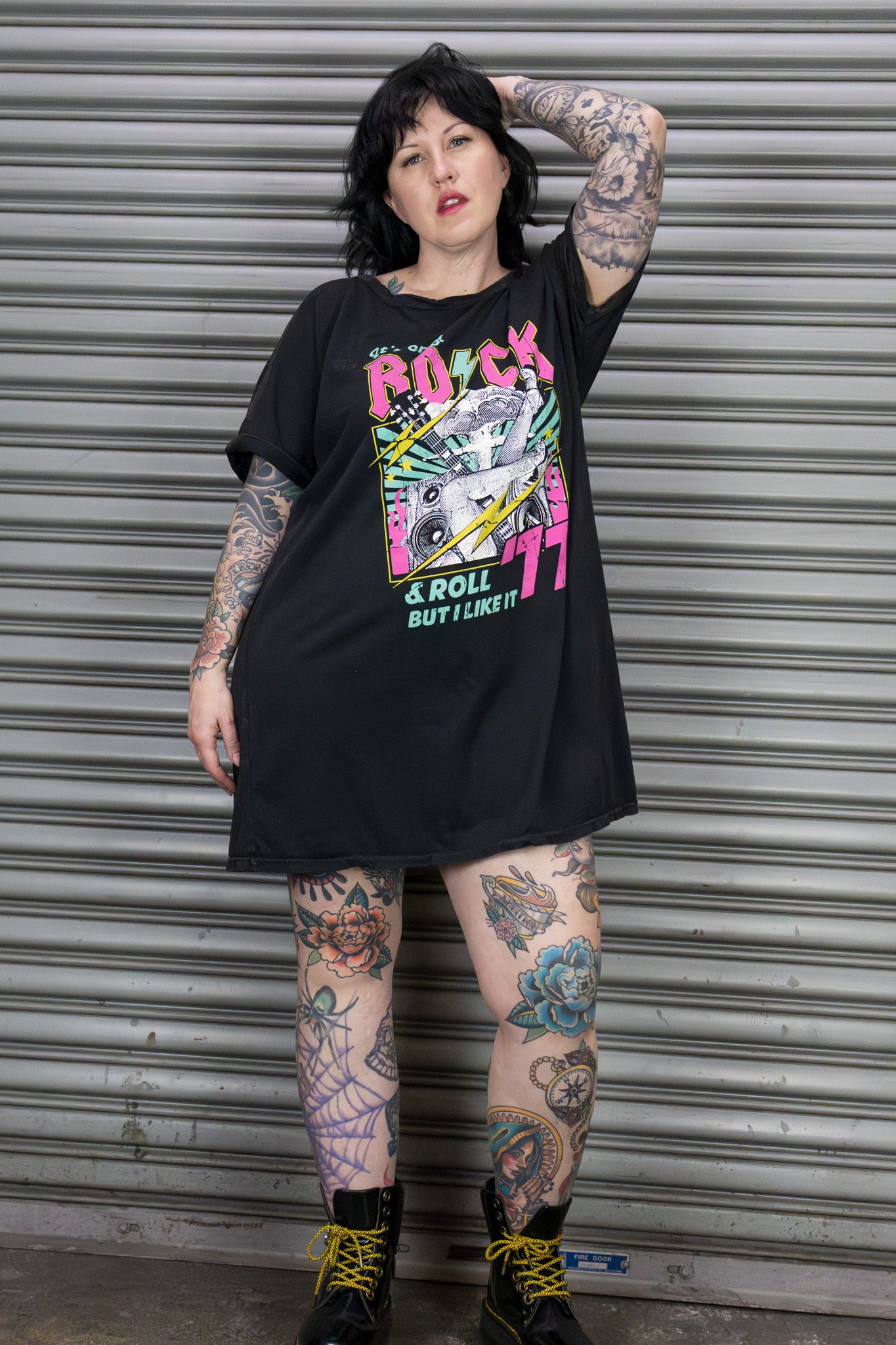 Person wearing a black oversized t-shirt with colorful graphics and text, standing in front of a metal shutter.