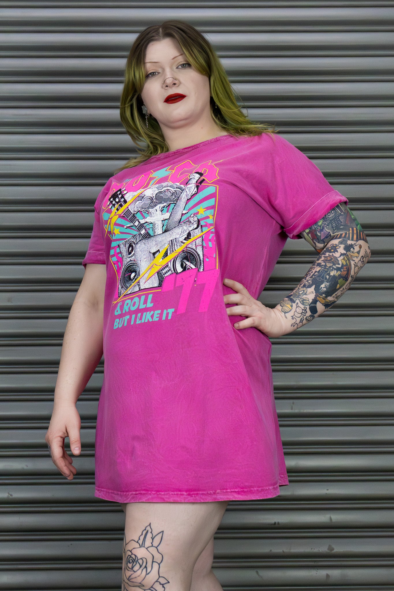 Person wearing a bright pink t-shirt with graphic design and text, standing against a metallic shutter background.