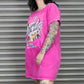 Person wearing a bright pink oversized t-shirt with a graphic design in front of a metallic shutter.