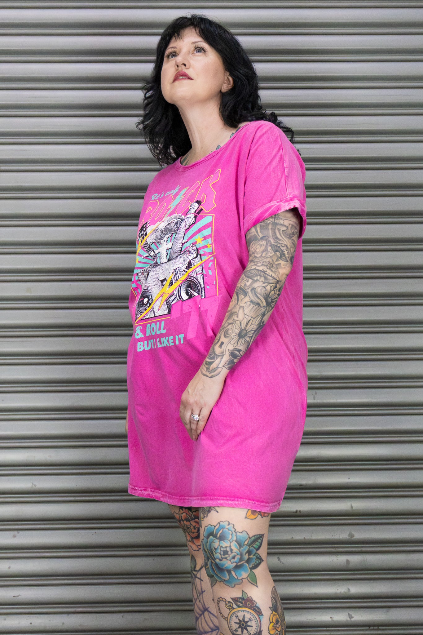 Person wearing a bright pink oversized t-shirt with a graphic design in front of a metallic shutter.