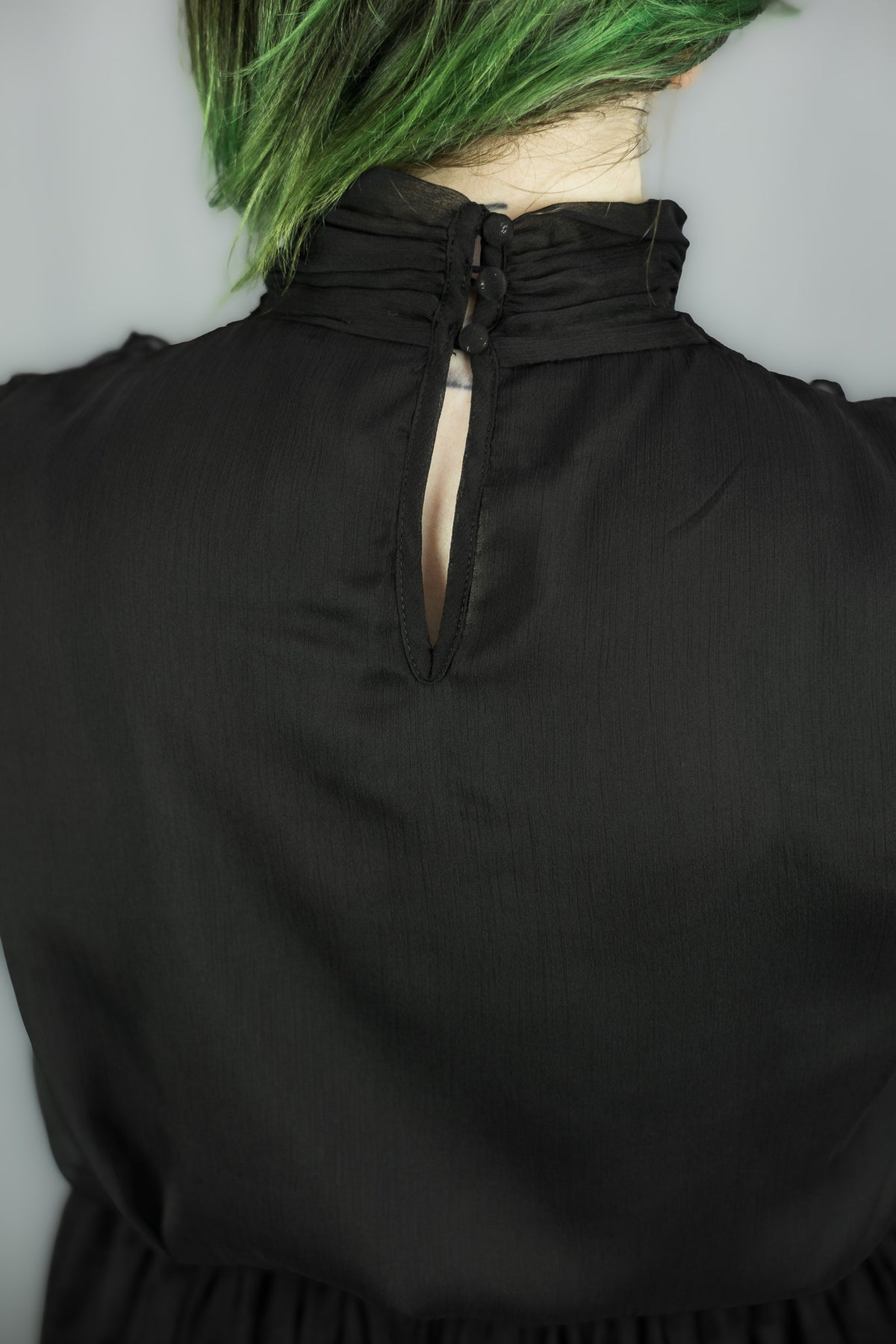 Close-up back view of a black dress on a person, showing a gathered high neck with a keyhole cutout and green hair visible at the top against a light gray background.